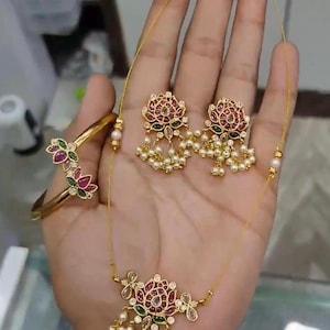 May include: A gold-coloured jewellery set comprising a necklace, earrings, and a bracelet. The floral designs incorporate red, green, and clear gemstones, with dangling faux pearls. The bracelet and necklace have a floral design. The earrings are a matching floral design.