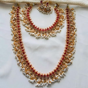 Puede incluir: Un conjunto de joyas de tres piezas que incluye un collar, un collar más largo y pendientes a juego. Las joyas son de color dorado con piedras preciosas rojas y detalles blancos similares a perlas. El conjunto se muestra sobre una superficie blanca.