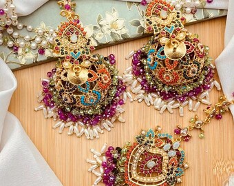 May include: An elaborate gold-coloured jewellery set, including a headpiece and earrings. The pieces are decorated with colourful gemstones in red, purple, green, and blue, and white pearl accents. The jewellery is displayed on a wooden surface.