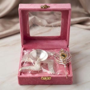 Puede incluir: Una caja de terciopelo rosa con tapa transparente contiene una lámpara diya plateada en forma de esvástica y un adorno de pluma de pavo real plateado decorado con pequeños cristales transparentes, envueltos en plástico.
