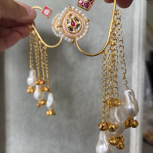 May include: Gold-toned ear cuff with a central teardrop-shaped design, adorned with pearls and gemstones. The cuff features dangling chains with pearl and gold bead accents, and small square accents with red gemstones.