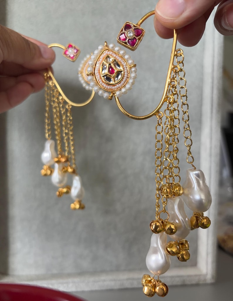 May include: Gold-toned ear cuff with a central teardrop-shaped design, adorned with pearls and gemstones. The cuff features dangling chains with pearl and gold bead accents, and small square accents with red gemstones.