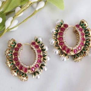 May include: A pair of gold-toned hoop earrings with a floral design. Each earring features a row of red gemstones, white pearls, and green and gold accents. The earrings are set against a white background.