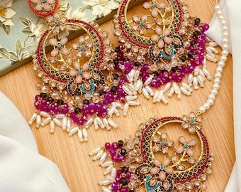 May include: Elaborate gold earrings with a floral motif. The earrings have a circular frame adorned with pink and clear gemstones, and small flower details. Clusters of purple and white beads dangle from the earrings. A matching headpiece is also present.