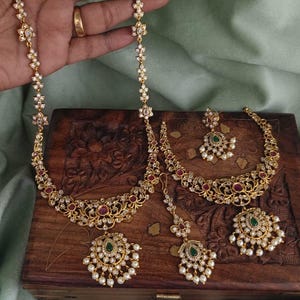 May include: A gold-toned jewelry set featuring a necklace, earrings, a bracelet, and a headpiece. The pieces are adorned with white gemstones, red accents, and a central green stone, set against a wooden box.