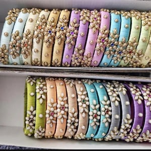 Peut inclure: Rangées de bracelets colorés dans une boîte. Les bracelets sont enveloppés de fil de soie et ornés d'ornements floraux dorés et de petites pierres précieuses colorées. Les couleurs comprennent le bleu clair, le jaune, le violet, le rose et le vert.