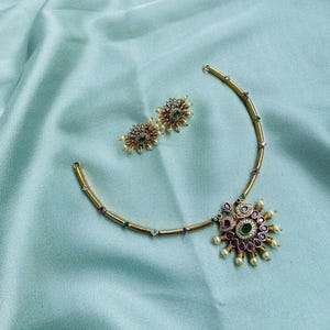 May include: A gold-toned necklace and matching earrings set. The necklace features a pendant with a central green stone, surrounded by red and white stones, and small pearls. The earrings have a similar design. The jewelry is set against a light blue fabric.