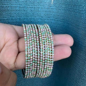 May include: A stack of gold-toned bangles adorned with alternating rows of pastel pink and mint green rhinestones. The bracelets are held against a teal background, showcasing their sparkling details. These accessories are perfect for adding a touch of glamour.