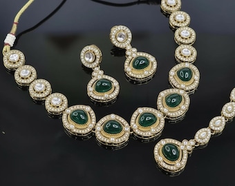 May include: A gold-toned jewelry set featuring a necklace and earrings. The necklace and earrings have emerald-green center stones surrounded by clear, sparkling accents. The set is displayed on a black surface.