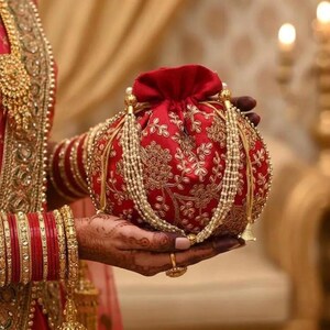 May include: A red, round pouch with gold floral embroidery and a drawstring closure. The pouch is adorned with beaded tassels and held in a hand with gold bangles and a ring. The background is blurred, suggesting a festive occasion.