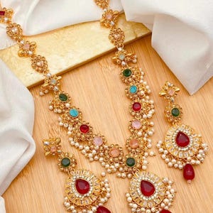May include: Gold-coloured necklace and earring set with colourful gemstones and faux pearls. The necklace and earrings feature a floral design with red, green, blue, and pink stones. The earrings have a large, teardrop-shaped red stone.