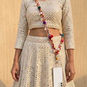 May include: A beige two-piece outfit featuring a cropped top and a long skirt, both adorned with intricate mirror work embroidery. A colorful braided crossbody phone strap with small pom-poms and white seashells is attached to a white smartphone.