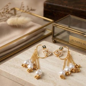 May include: A pair of gold-toned earrings with pearl and bead accents. The earrings feature a unique design with dangling chains and a square top with black and white details. The earrings are displayed on a marble surface.