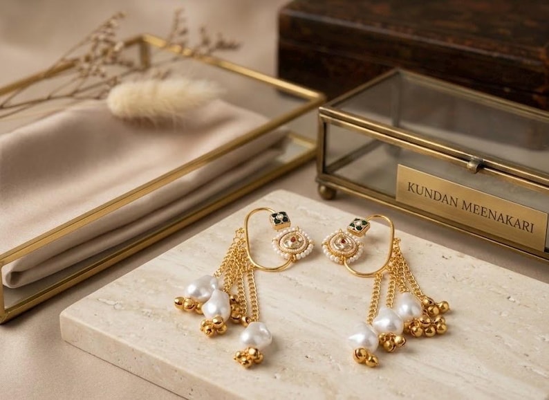 May include: A pair of gold-toned earrings with pearl and bead accents. The earrings feature a unique design with dangling chains and a square top with black and white details. The earrings are displayed on a marble surface.