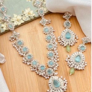 May include: A silver-toned necklace and matching earring set featuring light blue, oval-shaped stones and small white beads. The necklace has a central teardrop-shaped pendant, while the earrings feature a teardrop stone and a dangling green bead.