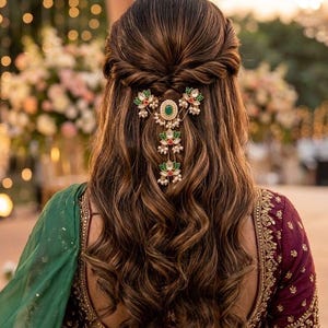 Könnte beinhalten: Ein Haarschmuck mit einem zentralen grünen Stein, umgeben von weißen Perlen und kleinen roten Akzenten, mit drei hängenden blütenförmigen Anhängern. Das Schmuckstück ist in einer halb hochgesteckten Frisur mit Locken und braunen Highlights befestigt.