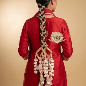 May include: A long, dark braided hairstyle adorned with an intricate gold-toned metal hair accessory featuring leaf-like patterns, dangling white cowrie shells, and small, multi-colored pastel tassels hanging from a geometric frame.