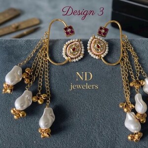 May include: Gold-toned earrings with pearl and chain accents. The earrings feature a square red gem at the top, a circular design with pearls, and dangling chains with white pearls and gold beads. The text "Design 3" and "ND jewelers" are visible.