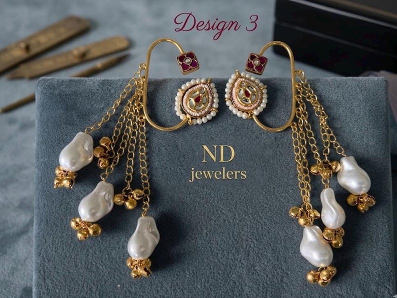 May include: Gold-toned earrings with pearl and chain accents. The earrings feature a square red gem at the top, a circular design with pearls, and dangling chains with white pearls and gold beads. The text "Design 3" and "ND jewelers" are visible.