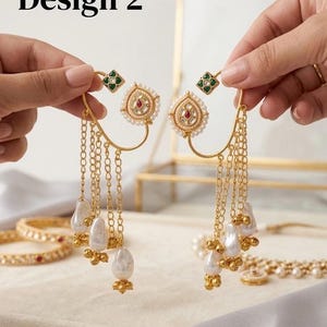 May include: Gold-toned earrings with a teardrop design, adorned with pearls and chains. The earrings feature a red gem at the center and green and white accents. The text "Design 2" is visible in the image.