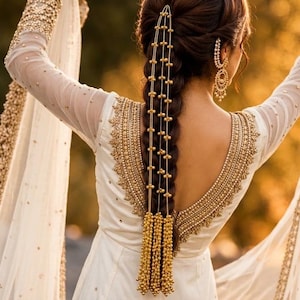 May include: A white long-sleeved dress featuring intricate gold embroidery along the neckline and cuffs, paired with a gold hair accessory consisting of multiple beaded chains draped over a braided hairstyle and matching gold drop earrings.