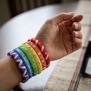 May include: A set of five stacked bangles in a rainbow color sequence of red, orange, yellow, green, blue, and purple. Each bangle is wrapped in fabric and decorated with metallic silver thread embroidery, small circular mirrors, and sequins.