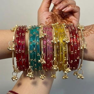 Könnte beinhalten: Eine Sammlung farbenfroher Armreifen in Rot, Türkis, Rosa, Gelb und Gold. Jeder Armreif ist mit kleinen, baumelnden goldenen Charms verziert. Die Armreifen sind an einem Handgelenk gestapelt und zeigen die lebendigen Farben und komplizierten Details.