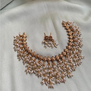 May include: A gold-coloured necklace and matching earrings set. The necklace features square-shaped gold links connected by strands of small, white, teardrop-shaped beads. The earrings mirror the design, creating a cohesive jewellery set.