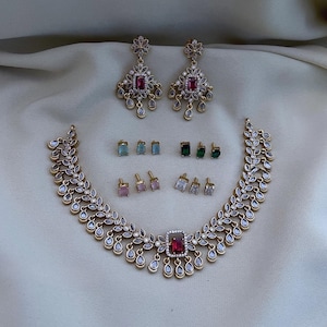 Puede incluir: Una colección de joyas doradas, que incluye un collar, pendientes y pendientes de botón. El collar y los pendientes presentan piedras preciosas rojas y transparentes. Los pendientes de botón vienen en varios colores, incluyendo rosa, verde y azul.
