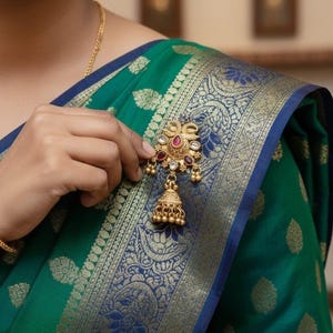 May include: A gold-toned brooch with a peacock design, featuring red and clear gemstones, and a dangling bell-shaped charm. The brooch is set against a teal and blue patterned fabric.