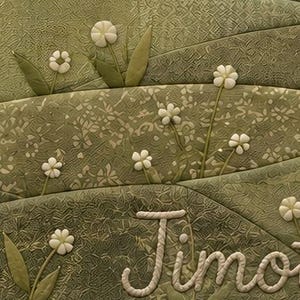 May include: A quilted textile featuring layered sage green fabric panels with textured patterns. The design includes three-dimensional white fabric flowers with green stems and leaves, and the name Timo embroidered in white rope-style lettering.