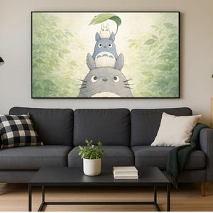 May include: A framed print of Totoro characters stacked on top of each other, with a green leaf held above. The artwork hangs above a dark gray sofa with pillows, a black coffee table, and a wooden bookshelf with books and a potted plant.