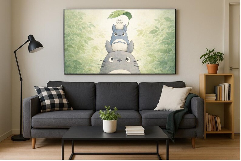 May include: A framed print of Totoro characters stacked on top of each other, with a green leaf held above. The artwork hangs above a dark gray sofa with pillows, a black coffee table, and a wooden bookshelf with books and a potted plant.