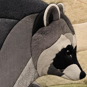 May include: A fabric quilt block featuring a stylised raccoon head constructed from various shades of grey, black, and white textured cotton fabrics. The design includes a dark nose, a round black eye, and distinct facial markings in white.