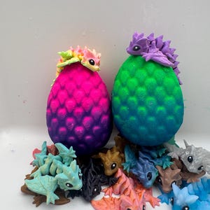 May include: Two large, textured, egg-shaped figures in vibrant pink-to-purple and green-to-blue gradients, each topped with a small dragon figurine. Surrounding the base are several smaller, multi-coloured articulated dragon figures in various poses.