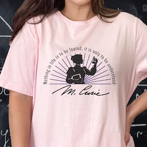 Könnte beinhalten: Ein hellrosa T-Shirt mit kurzen Ärmeln und einer schwarzen Grafik von Marie Curie, die ein Reagenzglas hält. Der Text "Nothing in life is to be feared, it is only to be understood" steht in einem Bogen darüber, "M. Curie" in Schreibschrift.