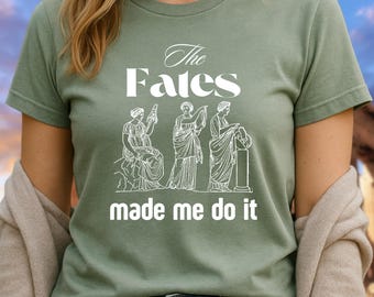 May include: Sage green t-shirt with the text "The Fates made me do it" and a graphic of three classical figures. The shirt is a short-sleeved crew neck. The background shows a building with columns.