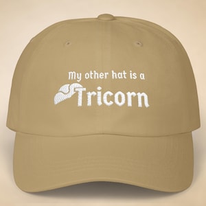 May include: A tan baseball cap with the embroidered phrase "My other hat is a Tricorn" in white. The hat has a curved brim and a button on top. The text is in a serif font, and the tricorn hat graphic is also white.