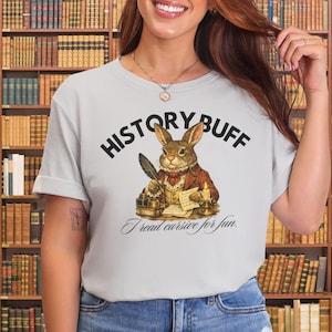 May include: A light grey t-shirt featuring a graphic of a rabbit dressed in a vintage waistcoat and cravat, writing with a quill at a desk. The text reads HISTORY BUFF in an arch above and I read cursive for fun in script below the illustration.