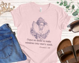 Pode incluir: Camiseta rosa claro com um retrato da rainha Elizabeth I e a citação «Não tenho nenhum desejo de fazer janelas nas almas dos homens». A imagem inclui jeans azuis, óculos de sol e sandálias marrons.