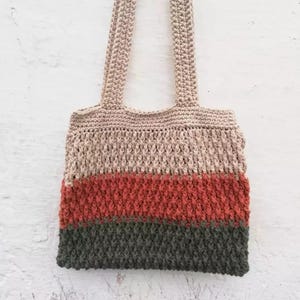 May include: A handmade crochet tote bag with a textured stitch pattern. The bag features horizontal stripes in beige, rust, and dark gray. The handles are a matching beige color. The bag is hanging against a white background.