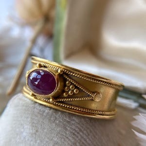 May include: A gold ring with an oval, deep red gemstone. The ring has an ornate design with decorative elements and a textured band. The ring is displayed on a soft, light-colored surface.