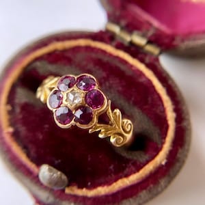 Peut inclure: Une bague en or ornée d'un motif floral composé de six rubis rouge foncé entourant un diamant blanc central. L'anneau présente des détails métalliques travaillés en forme de feuilles et est présenté dans un écrin doublé de velours rouge.