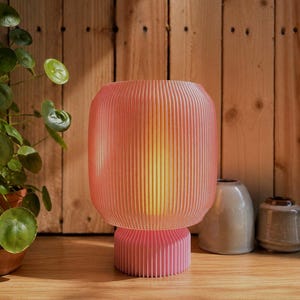 Könnte beinhalten: Eine Tischleuchte mit einem zylindrischen, gerippten Lampenschirm in einem sanften Korallenrosa, die auf einem passenden, kleineren, kreisförmigen gerippten Sockel ruht. Die Lampe strahlt ein warmes, goldenes Licht aus, das die Struktur betont.