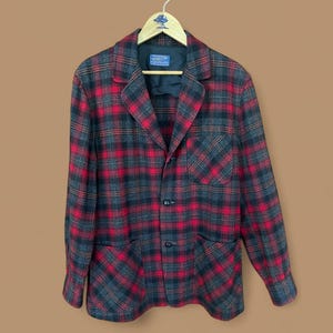 May include: A red, black, and gray plaid blazer with a notched collar, two front pockets, and three buttons. The blazer is hanging on a wooden hanger.