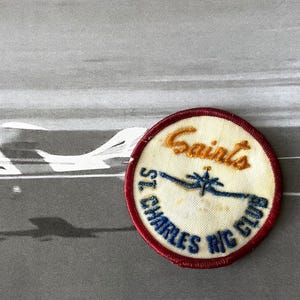 May include: A vintage patch with the text "Saints St. Charles R/C Club" in blue and orange embroidery. The patch is round with a red border. A small airplane is blurred in the background.
