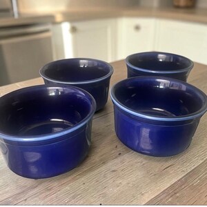 May include: A set of four deep, circular ceramic bowls in a solid, glossy cobalt blue finish. Each bowl features a smooth, rounded rim with a subtle, lighter blue accent line along the top edge, providing a consistent look across the collection.