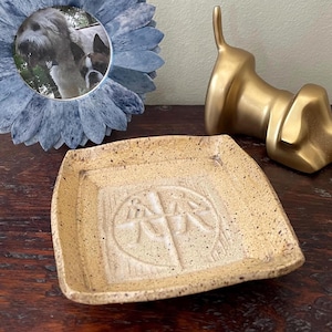 May include: A decorative arrangement featuring a light blue flower-shaped picture frame with a dog photo, a gold-colored dog sculpture, and a beige square ceramic dish with a textured surface and a central design.