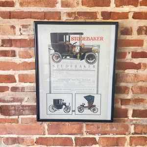 May include: Framed vintage advertisement for Studebaker automobiles. The advertisement features a detailed illustration of a classic car, with the Studebaker logo prominently displayed. Additional smaller illustrations of other models are included. The frame is black.