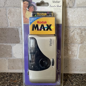 May include: A Vivitar Point 'n Shoot 35mm Camera Kit. The kit includes a silver and black camera, a box of Kodak MAX film, and Kodak alkaline batteries. The packaging features a photo of a woman and child.